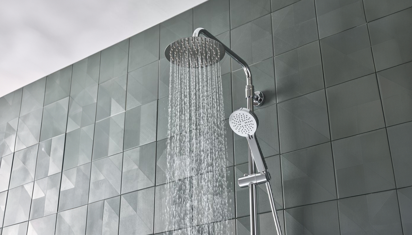 A modern shower with a large overhead showerhead and a handheld showerhead. The showerhead is mounted on a chrome pole against a wall of gray tiles with a geometric pattern. Water is flowing from the overhead showerhead