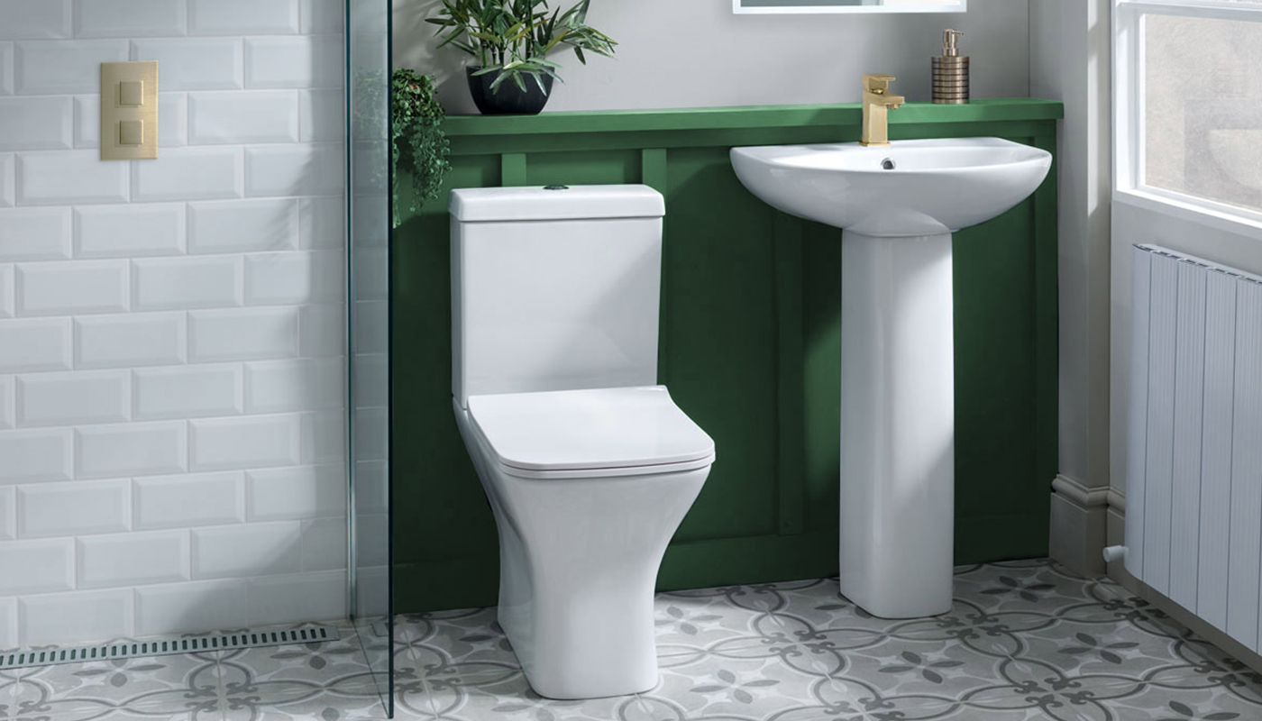 A modern bathroom with white subway tile walls, a white toilet with a dual flush mechanism, a white pedestal sink with a gold faucet, and a green accent wall with a shelf holding a plant. The floor is tiled with a patterned design in shades of gray