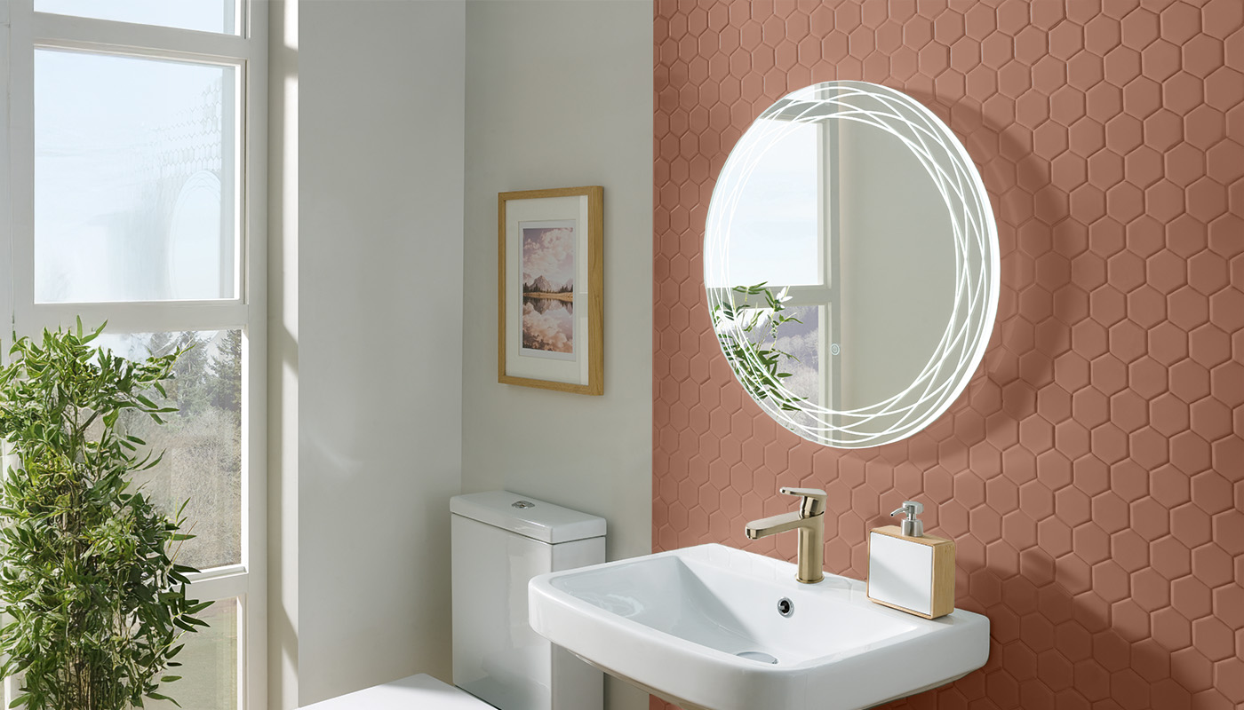 A bright and airy bathroom with a large window and a round illuminated mirror. The mirror has a white frame with a double ring of warm white LED lights. Below the mirror is a white ceramic sink and a gold faucet. The wall behind the mirror is covered in Mocha Mousse brown hexagonal mosaic tiles.